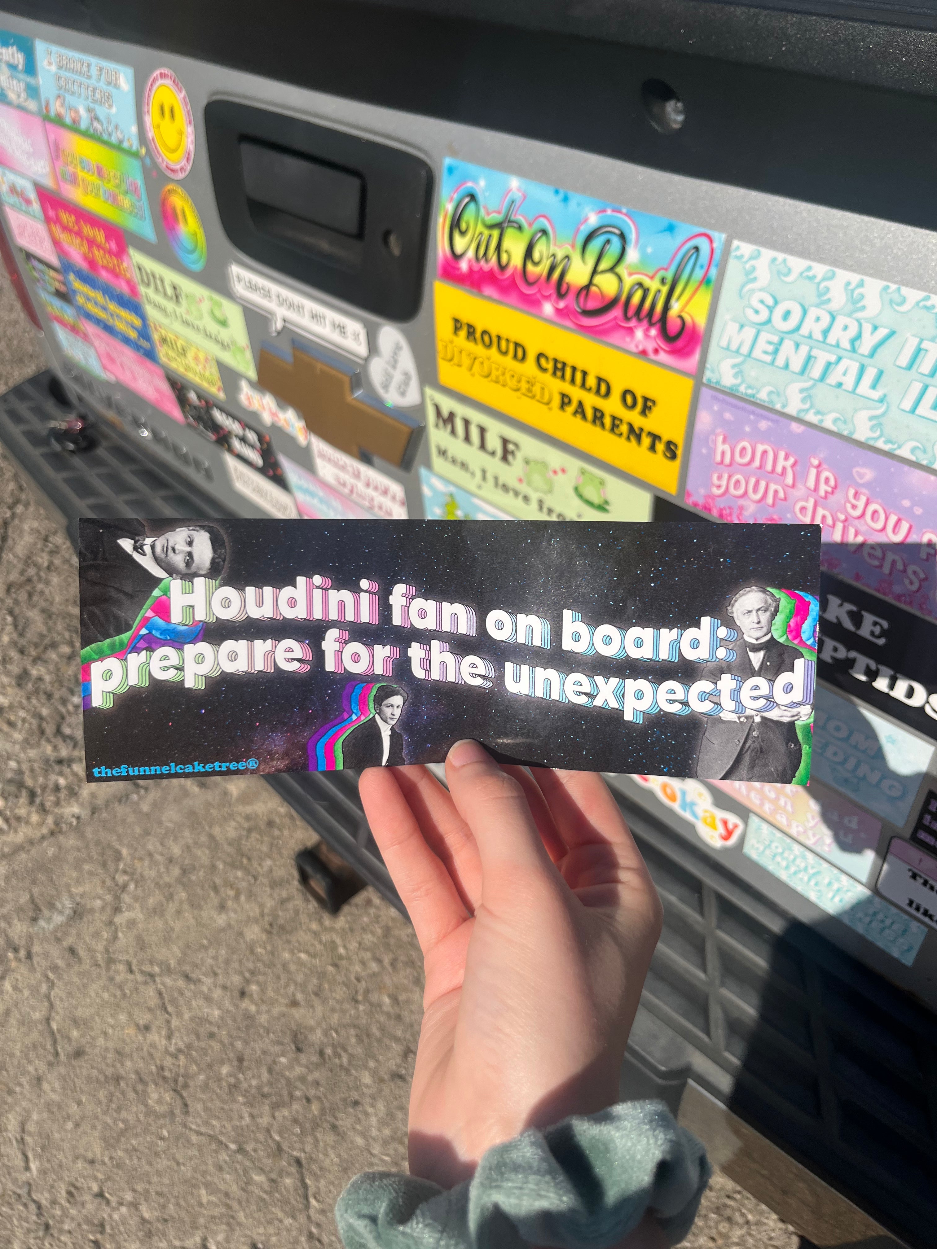 A hand holding a black bumper sticker with white text saying 'Houdini fan on board: prepare for the unexpected'.