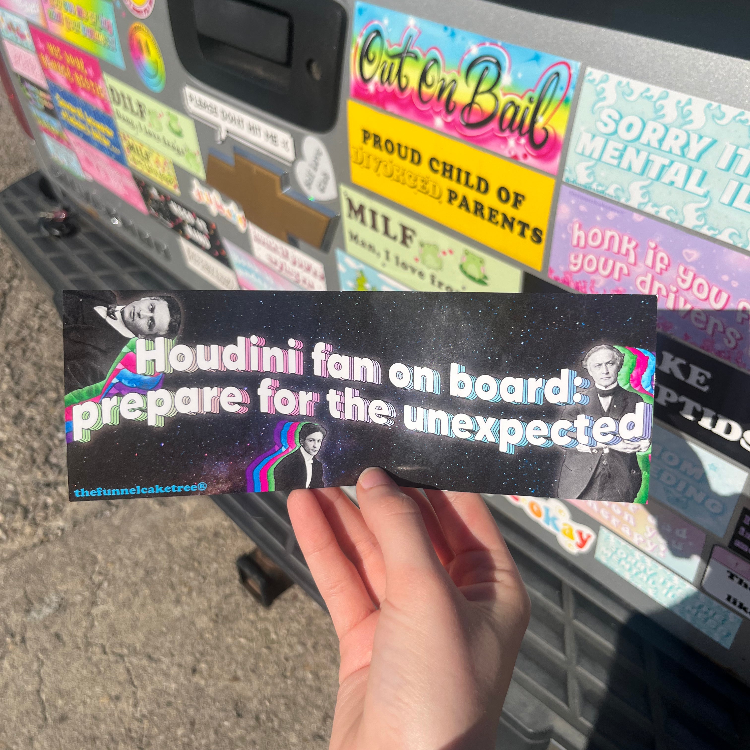 A hand holding a black bumper sticker with white text saying 'Houdini fan on board: prepare for the unexpected'.