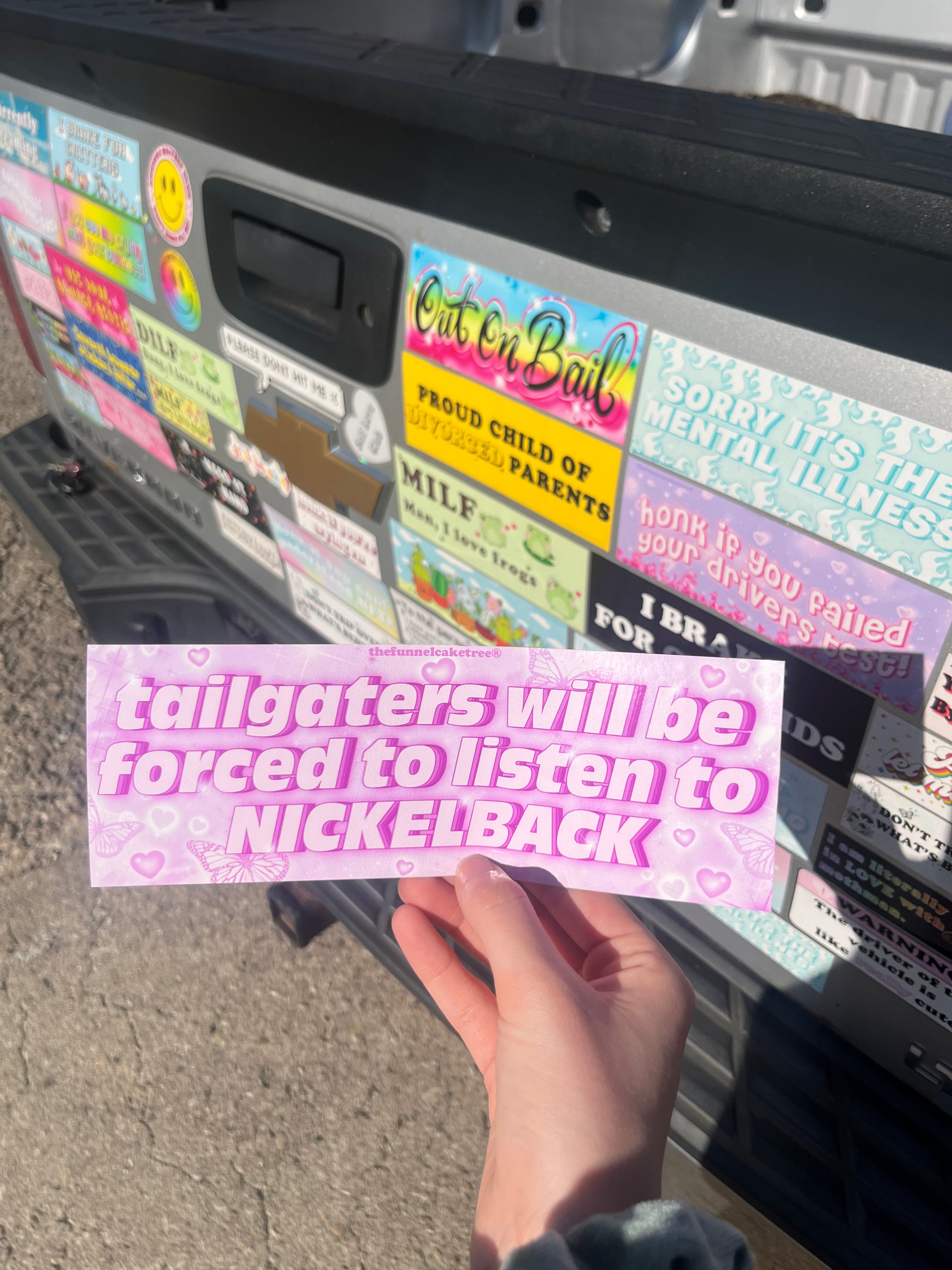 A hand holding a rectangular bumper sticker with the text 'Tailgaters - will be forced to listen to NICKELBACK' in pink color on a background showing various other bumper stickers.