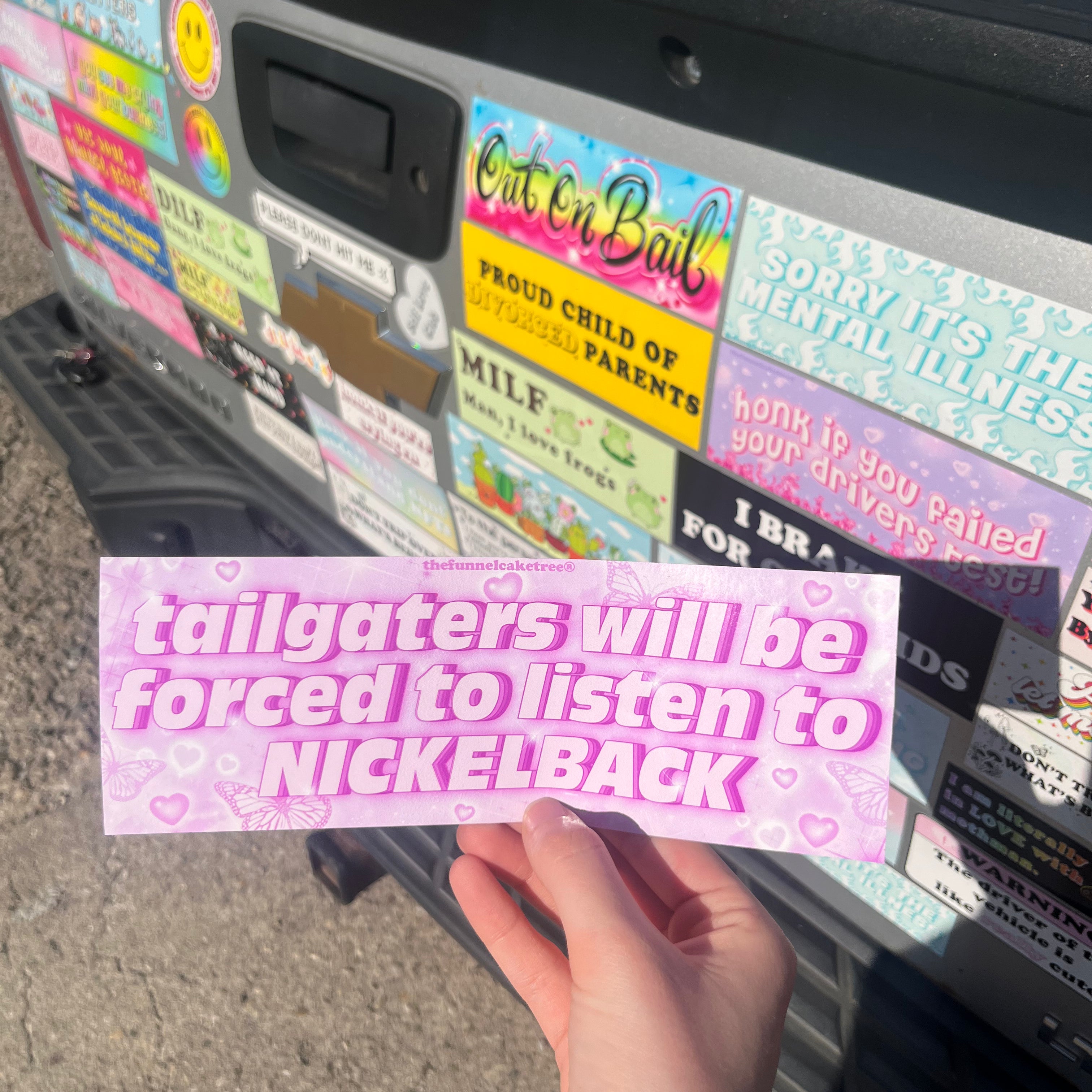 A hand holding a rectangular bumper sticker with the text 'Tailgaters - will be forced to listen to NICKELBACK' in pink color on a background showing various other bumper stickers.