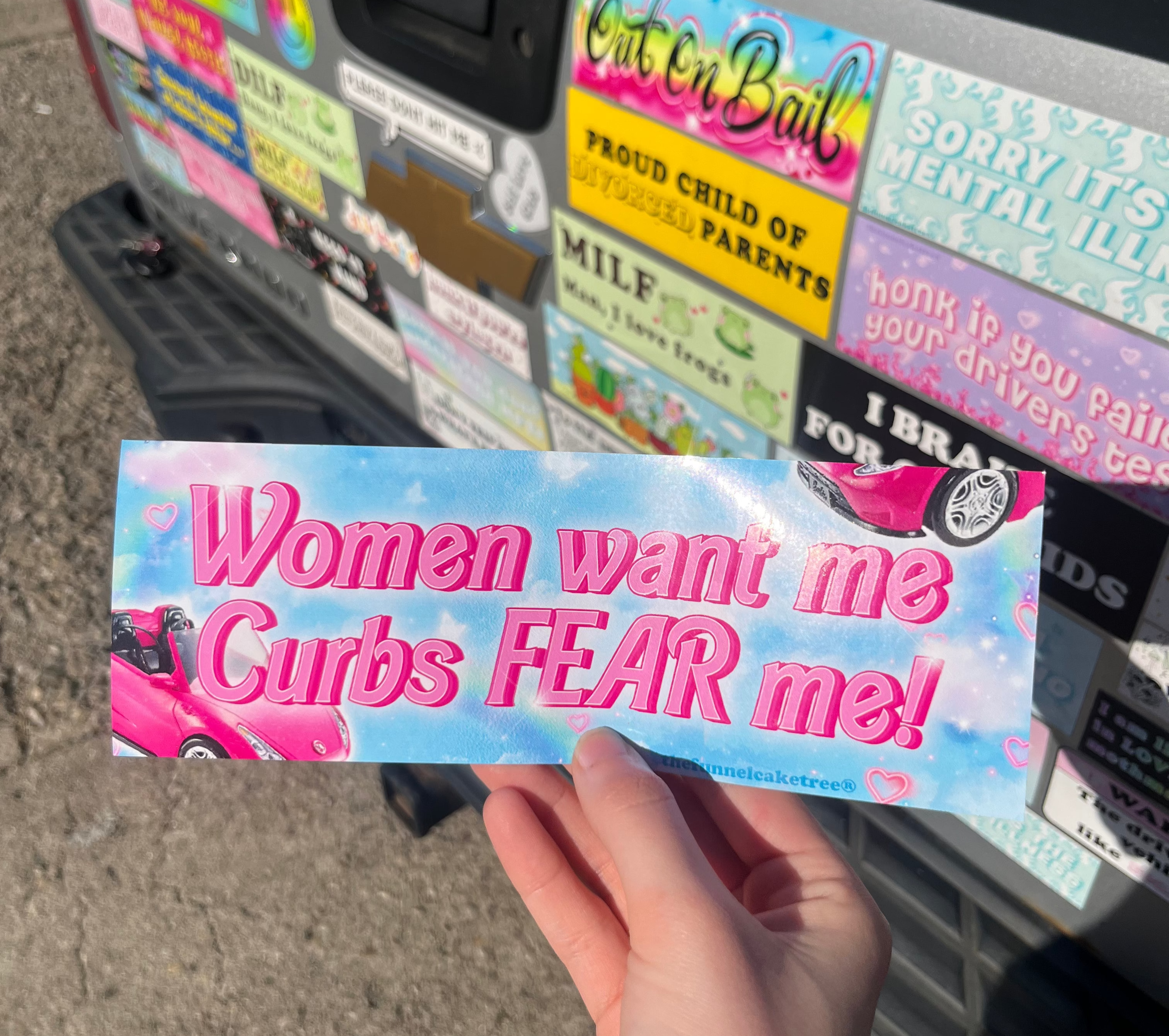 Izzy with The Funnel Cake Tree holding a bumper sticker with the text  Women want me curbs fear me.  Behind the sticker you see the tailgate of her truck that is covered in bumper stickers. 