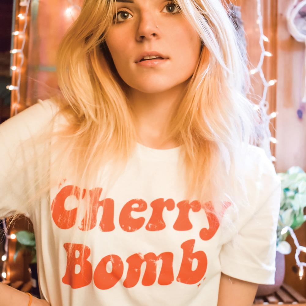 Woman wearing a white ‘Cherry Bomb’ graphic tee with bold red retro lettering, standing in a cozy room with string lights.