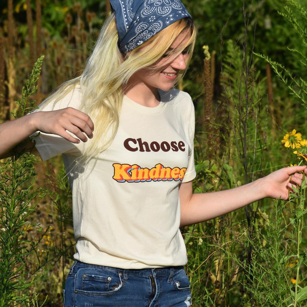 Smiling woman in a cream ‘Choose Kindness’ t-shirt and denim jeans walking through a wildflower field on a sunny day.