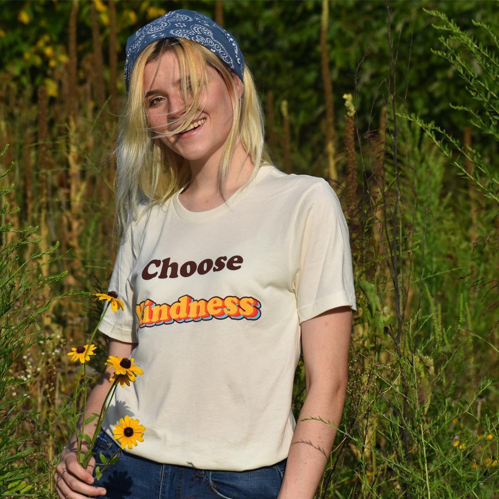 Woman wearing a cream ‘Choose Kindness’ graphic tee with bold retro lettering, standing in a sunny field holding yellow flowers.