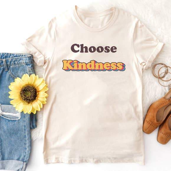 Flat lay of a cream ‘Choose Kindness’ graphic tee with retro orange and blue lettering, styled with denim jeans, sunflower, and tan flats.