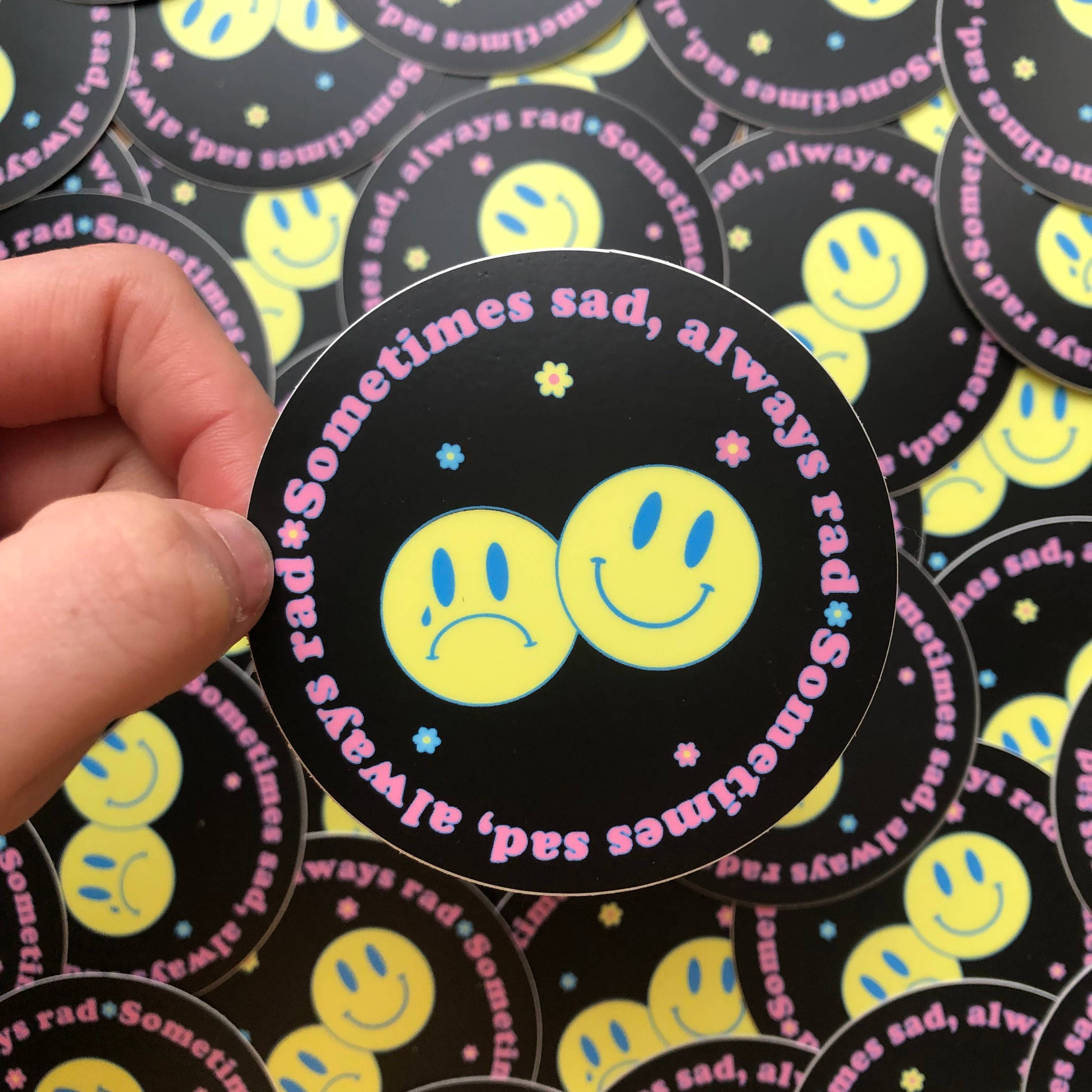 Round black sticker with two yellow smiley faces and the text Sometimes sad, always rad.