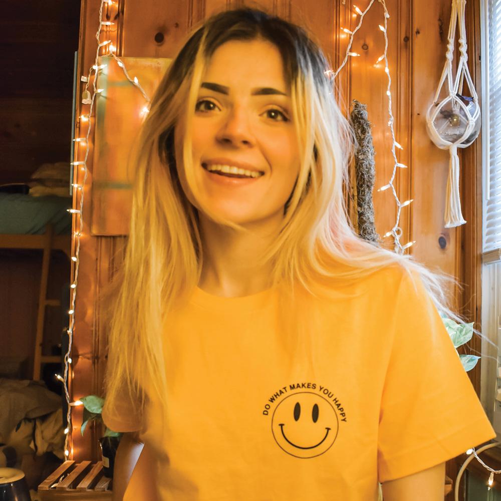Smiling person wearing yellow ‘Do What Makes You Happy’ t-shirt by Funnel Cake Tree with black smiley face print , retro positive vibes graphic tee.