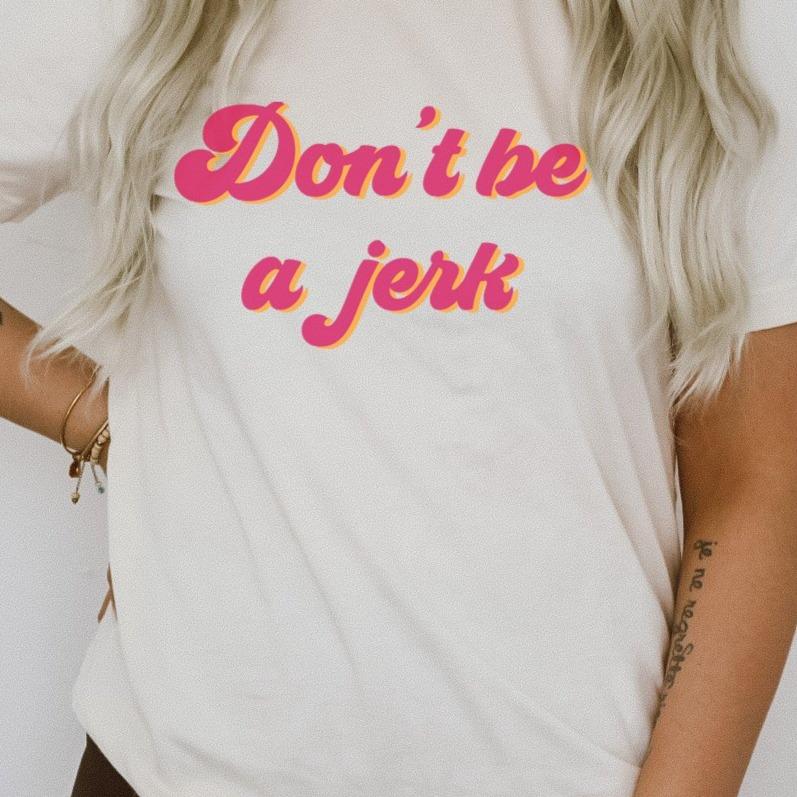 Woman wearing a natural color ‘Don’t Be a Jerk’ graphic tee with pink and yellow lettering, paired with casual wavy hair and accessories.