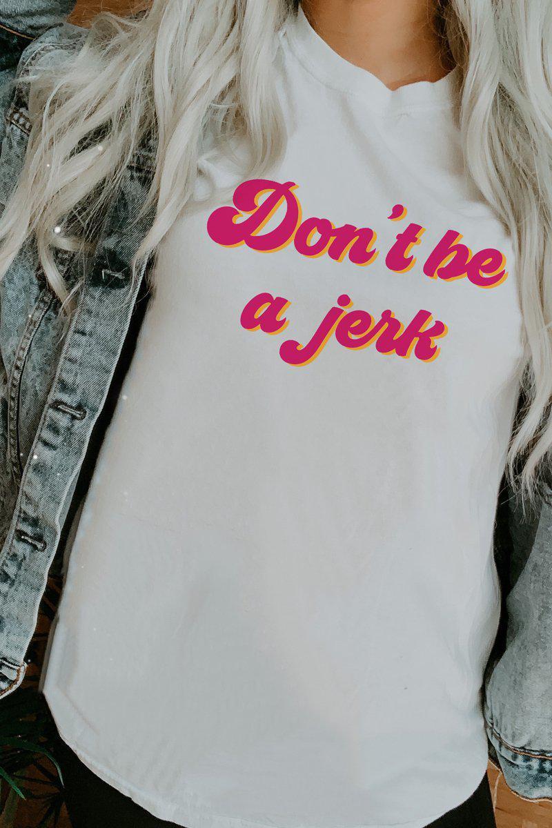 Woman wearing a white ‘Don’t Be a Jerk’ graphic tee with pink and yellow retro lettering, styled with a denim jacket.