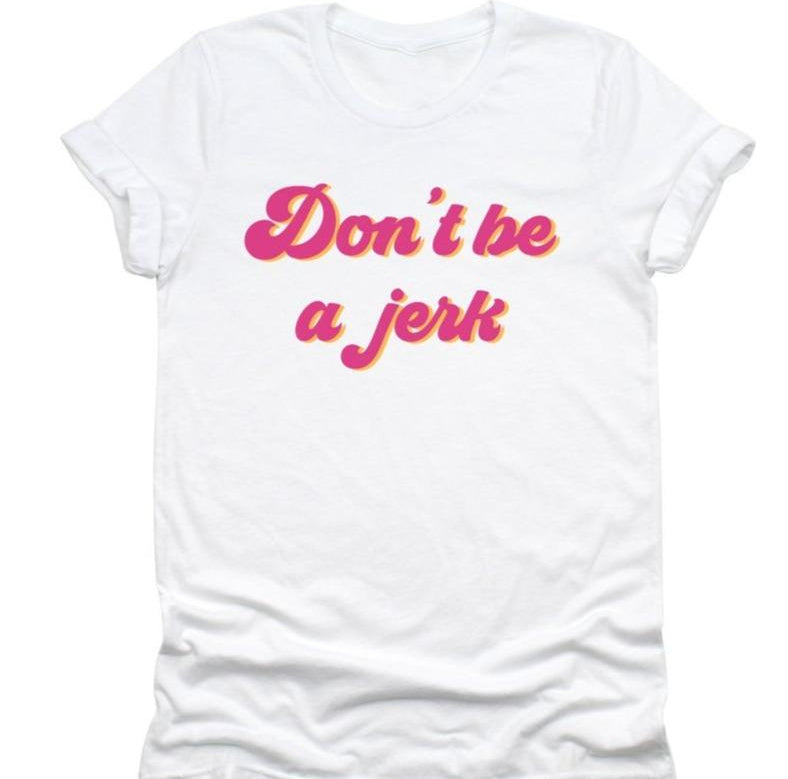 Flat lay of a white ‘Don’t Be a Jerk’ graphic tee featuring bold pink and yellow retro lettering, laid out neatly on a white background.