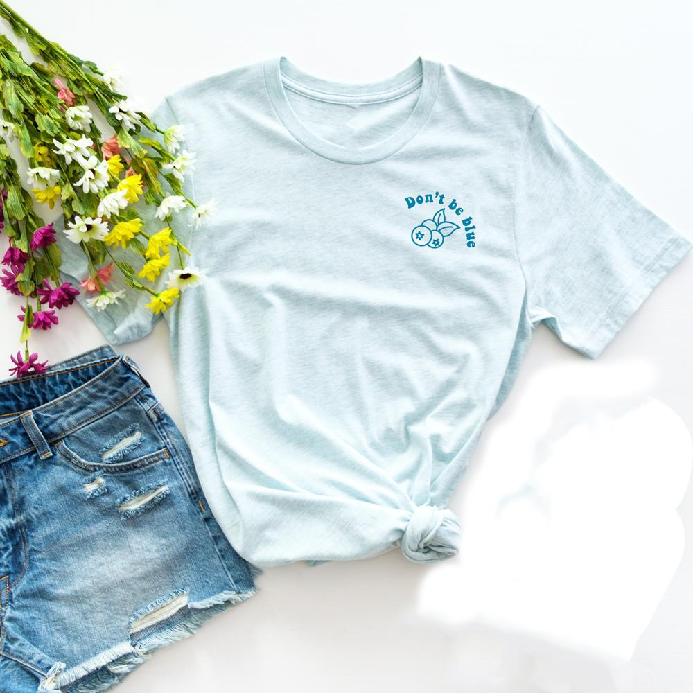 Light blue ‘Don’t Be Blue’ graphic tee by Funnel Cake Tree featuring a blueberry design and positive message, flat lay with denim shorts and colorful flowers.