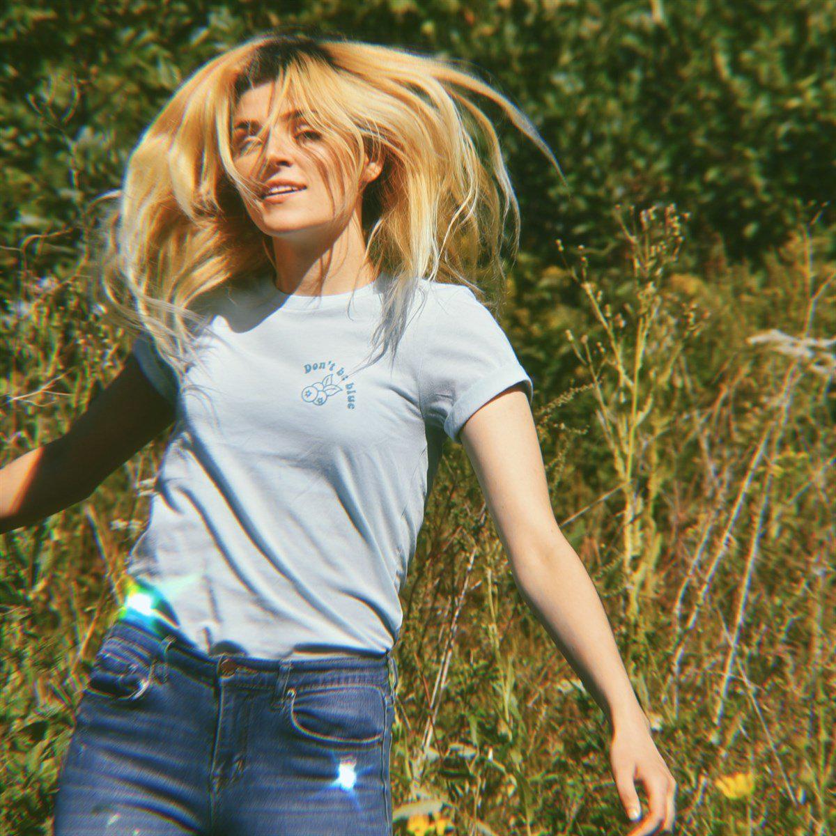 Light blue ‘Don’t Be Blue’ graphic tee by Funnel Cake Tree featuring a small blueberry design and uplifting message, worn outdoors in a sunny field