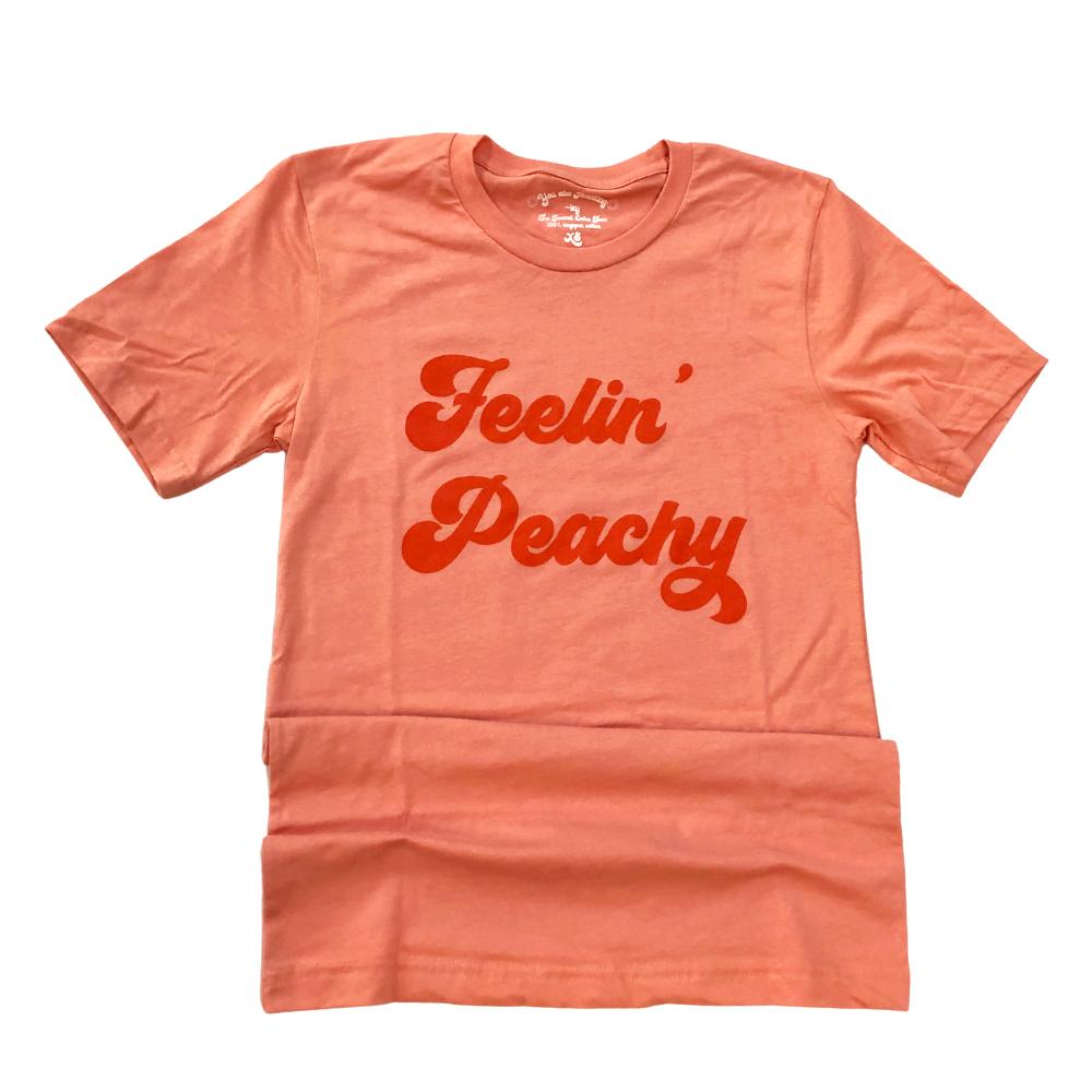 Feelin’ Peachy flat lay graphic tee by Funnel Cake Tree peach-colored unisex t-shirt with bold retro red lettering displayed on a flat surface.