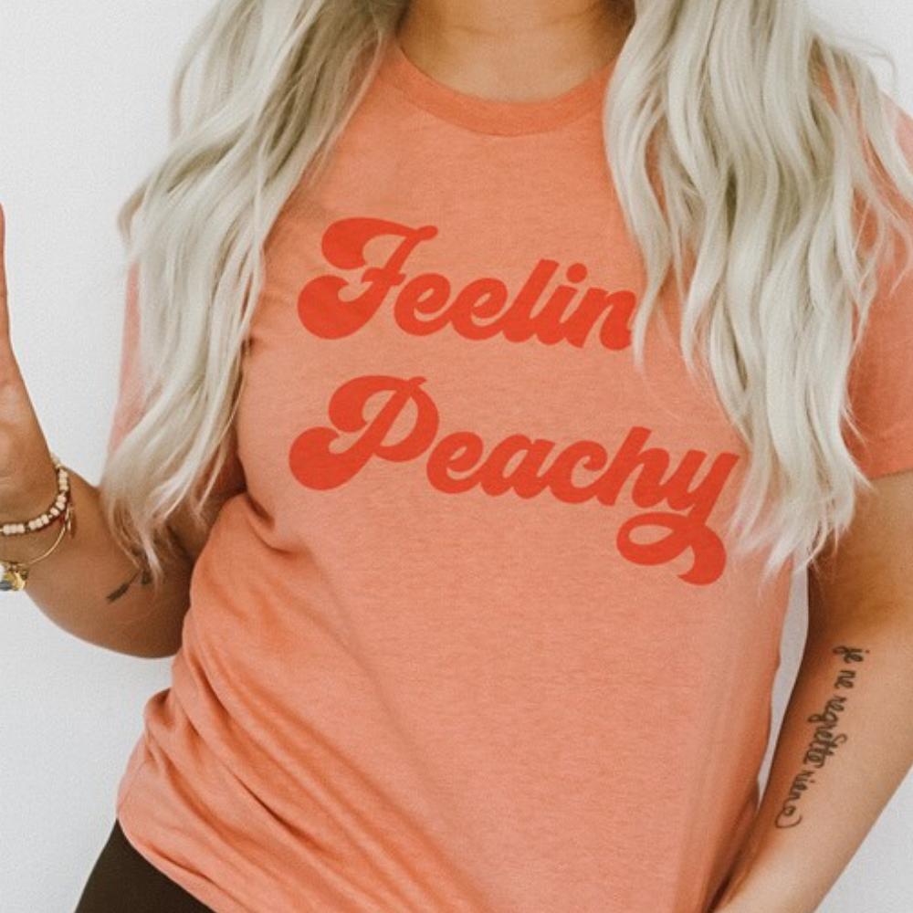Feelin’ Peachy retro graphic tee by Funnel Cake Tree model wearing peach-colored t-shirt with bold vintage red lettering, styled casually for a soft lifestyle look.