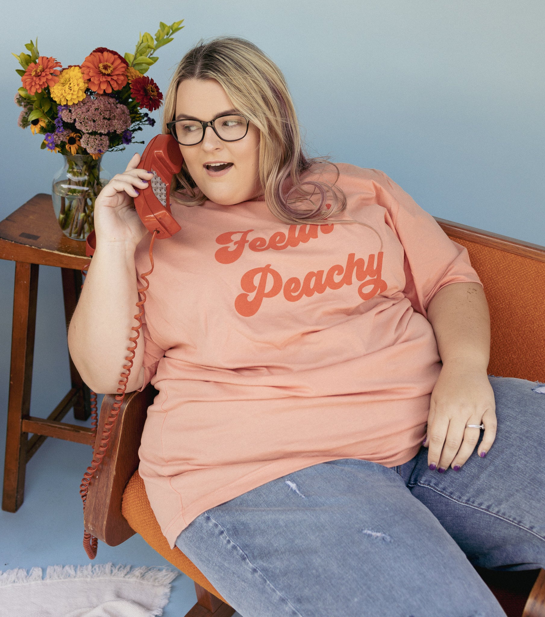 Feelin’ Peachy graphic t-shirt by Funnel Cake Tree model wearing peach-colored tee with bold retro red lettering, sitting on vintage orange chair and holding red phone for a fun, nostalgic lifestyle look.