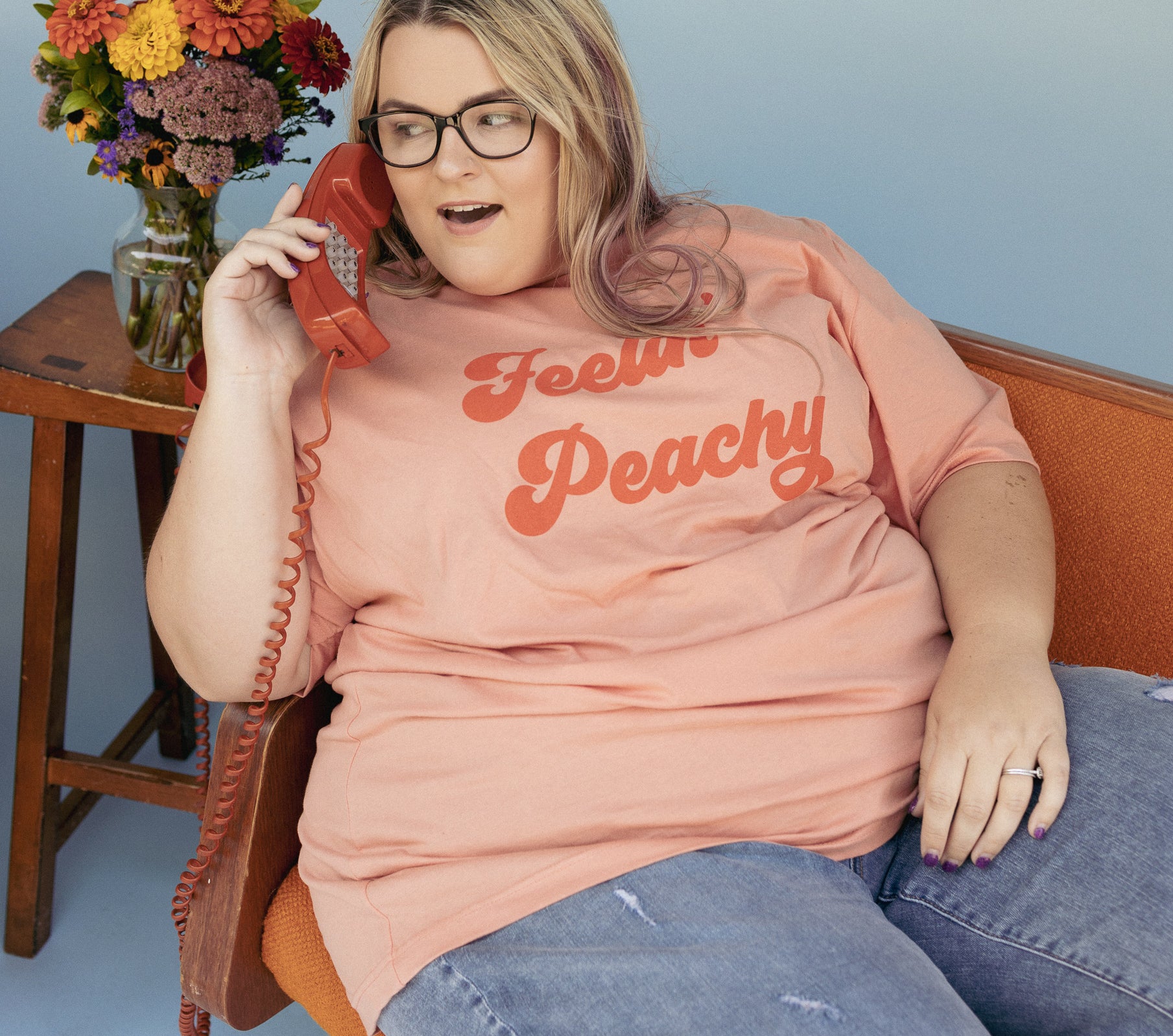 Feelin’ Peachy graphic t-shirt by Funnel Cake Tree model wearing peach-colored tee with bold retro red lettering, sitting on vintage orange chair and holding red phone for a fun, nostalgic lifestyle look.