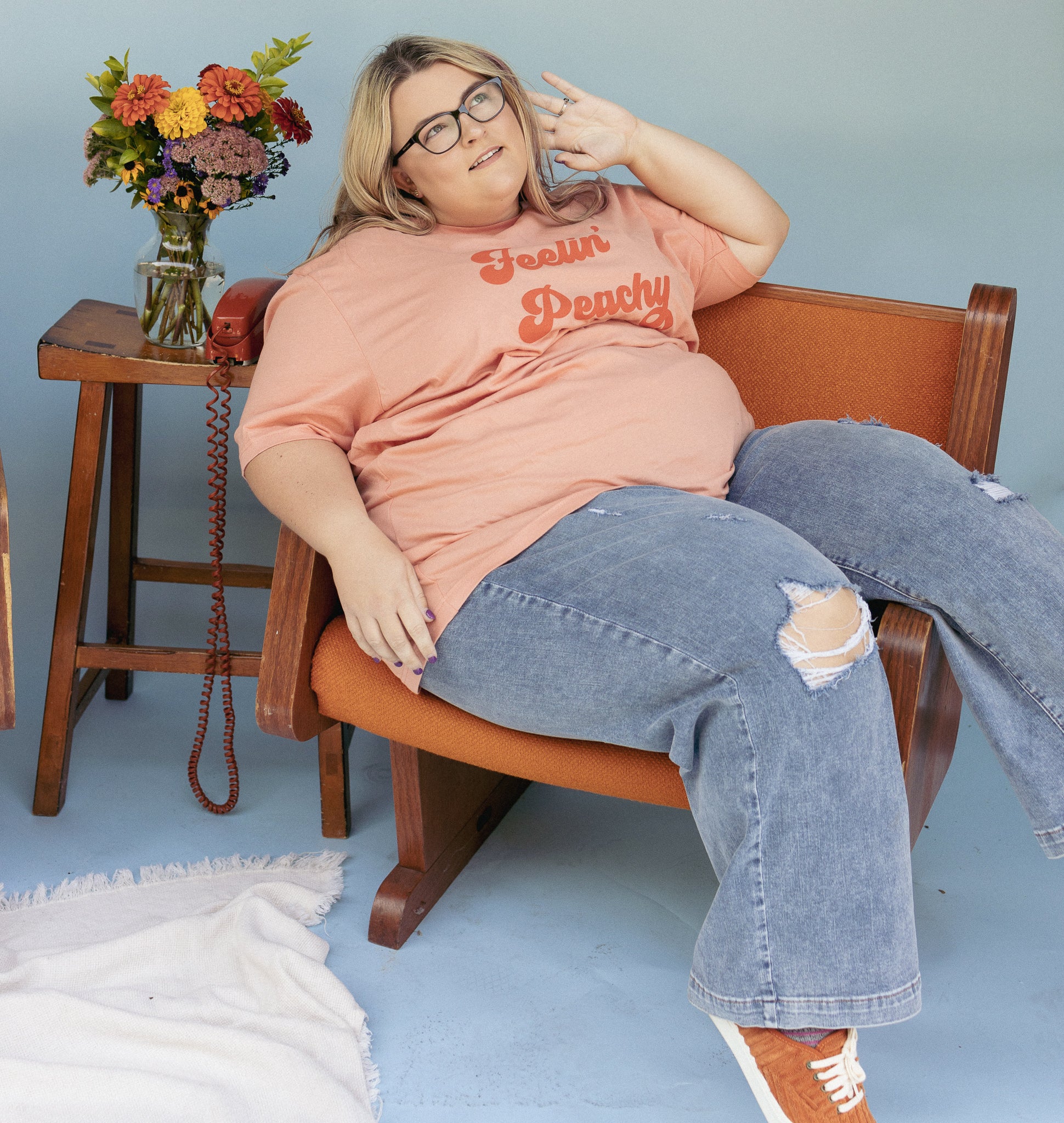 Feelin’ Peachy retro graphic tee by Funnel Cake Tree, lifestyle photo featuring model wearing peach-colored t-shirt with bold vintage lettering, seated on orange chair beside floral bouquet.