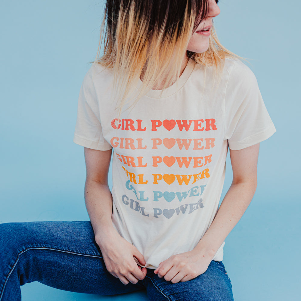 Person wearing a cream ‘Girl Power’ graphic tee with pastel rainbow lettering, styled with jeans against a blue background, photographed for Funnel Cake Tree.