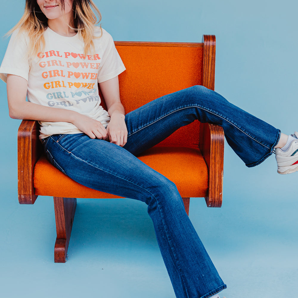 Person sitting on an orange chair wearing a cream ‘Girl Power’ graphic tee with pastel rainbow lettering, styled with jeans, photographed for Funnel Cake Tree.