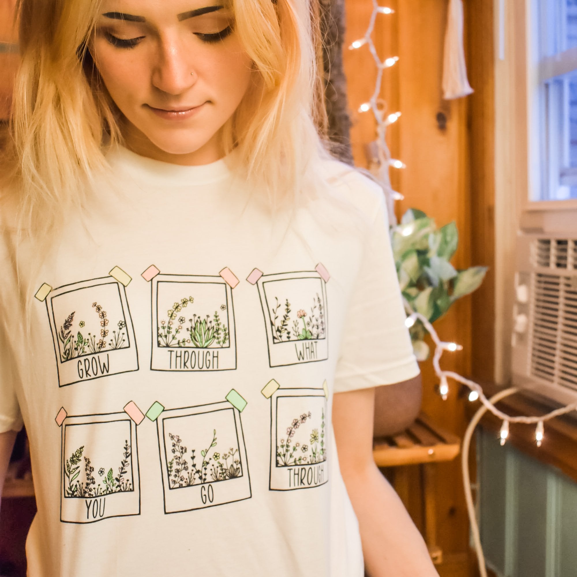 Grow Through What You Go Through lifestyle graphic tee by Funnel Cake Tree cream unisex t-shirt featuring hand-drawn floral frames with motivational lettering, modeled indoors with cozy string lights.