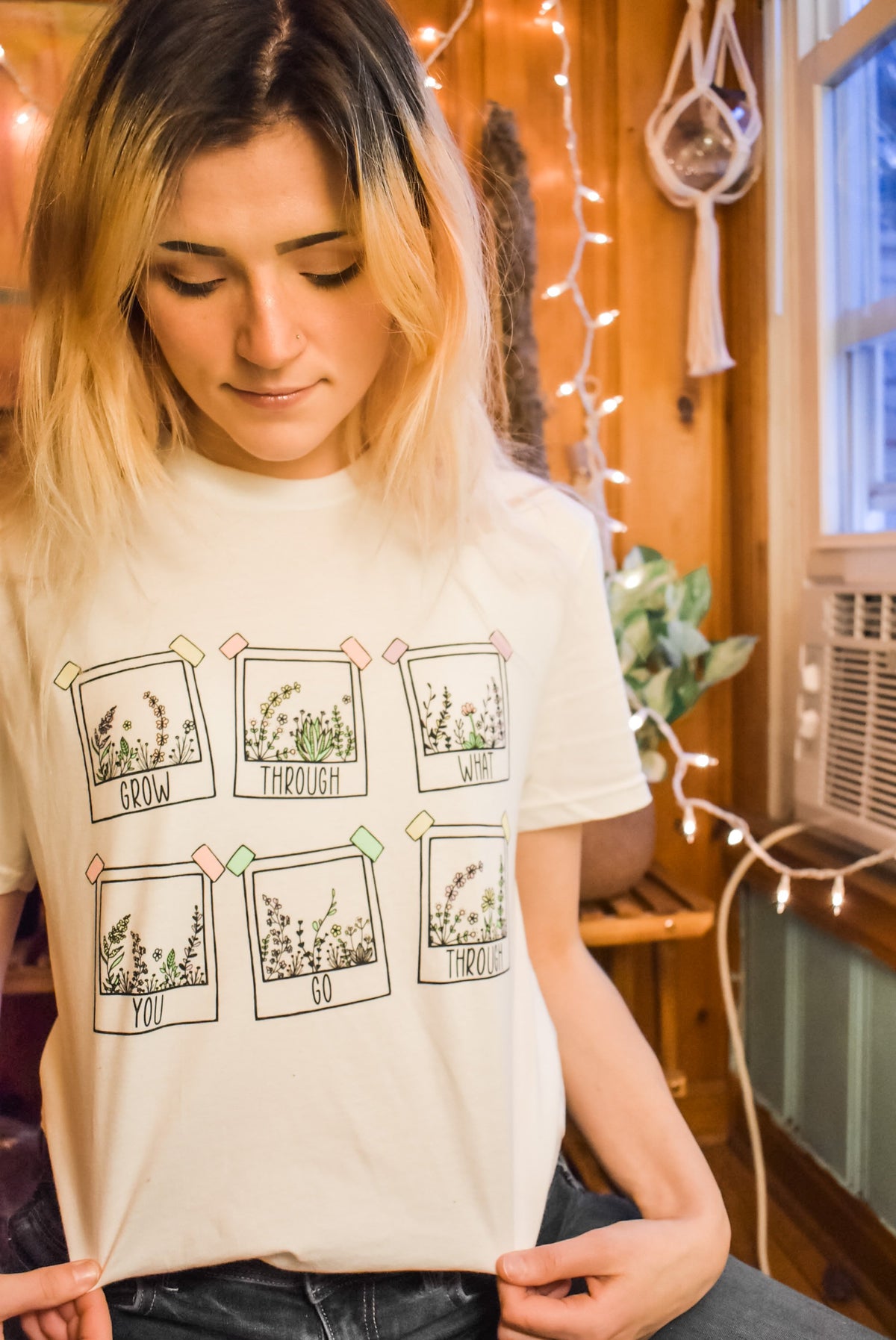 Grow Through What You Go Through lifestyle graphic tee by Funnel Cake Tree cream unisex t-shirt featuring hand-drawn floral frames with motivational lettering, modeled indoors with cozy string lights.