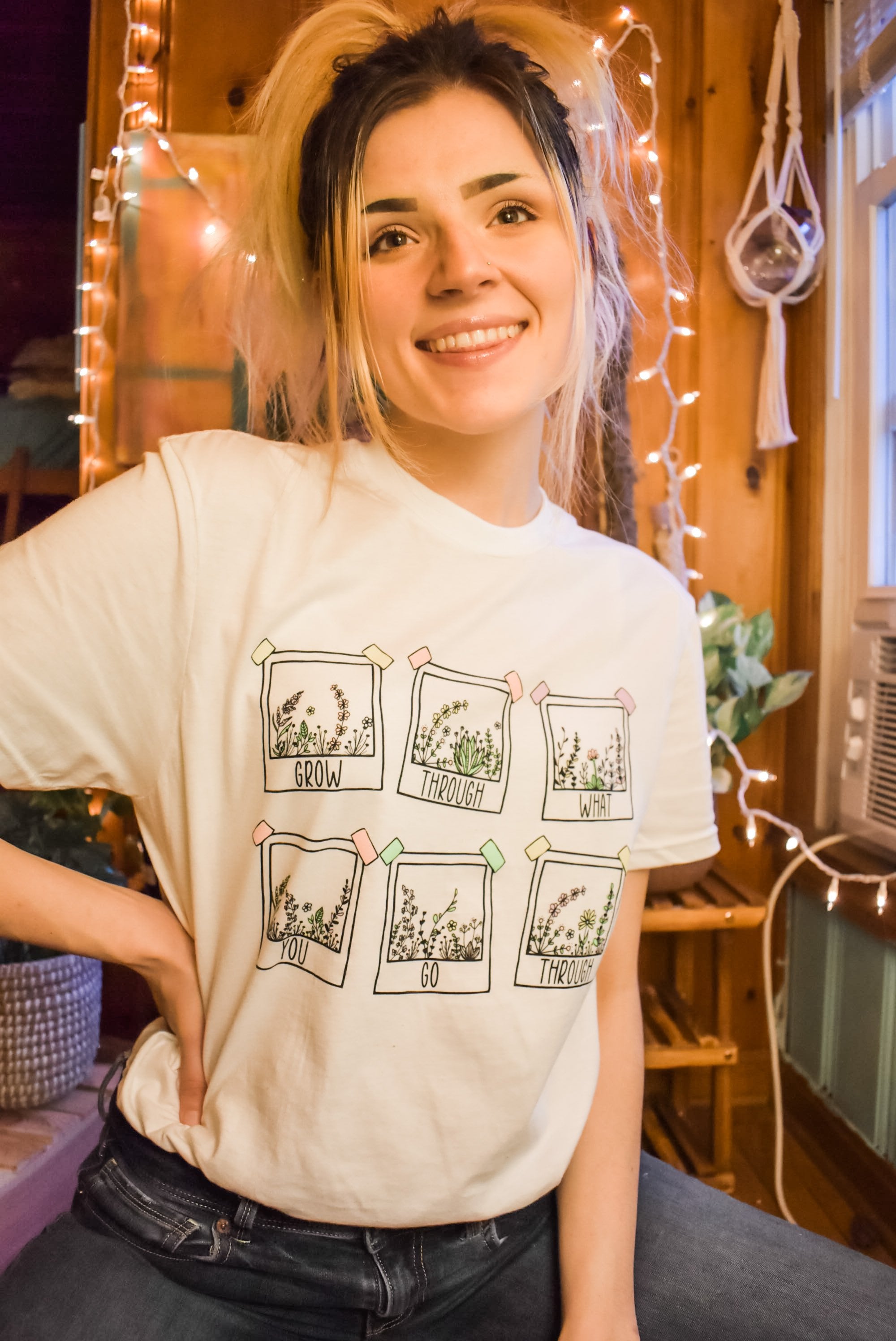 Grow Through What You Go Through positive message tee by Funnel Cake Tree cream unisex t-shirt featuring six floral polaroid-style frames and uplifting lettering, modeled indoors with string lights and a cozy vibe.