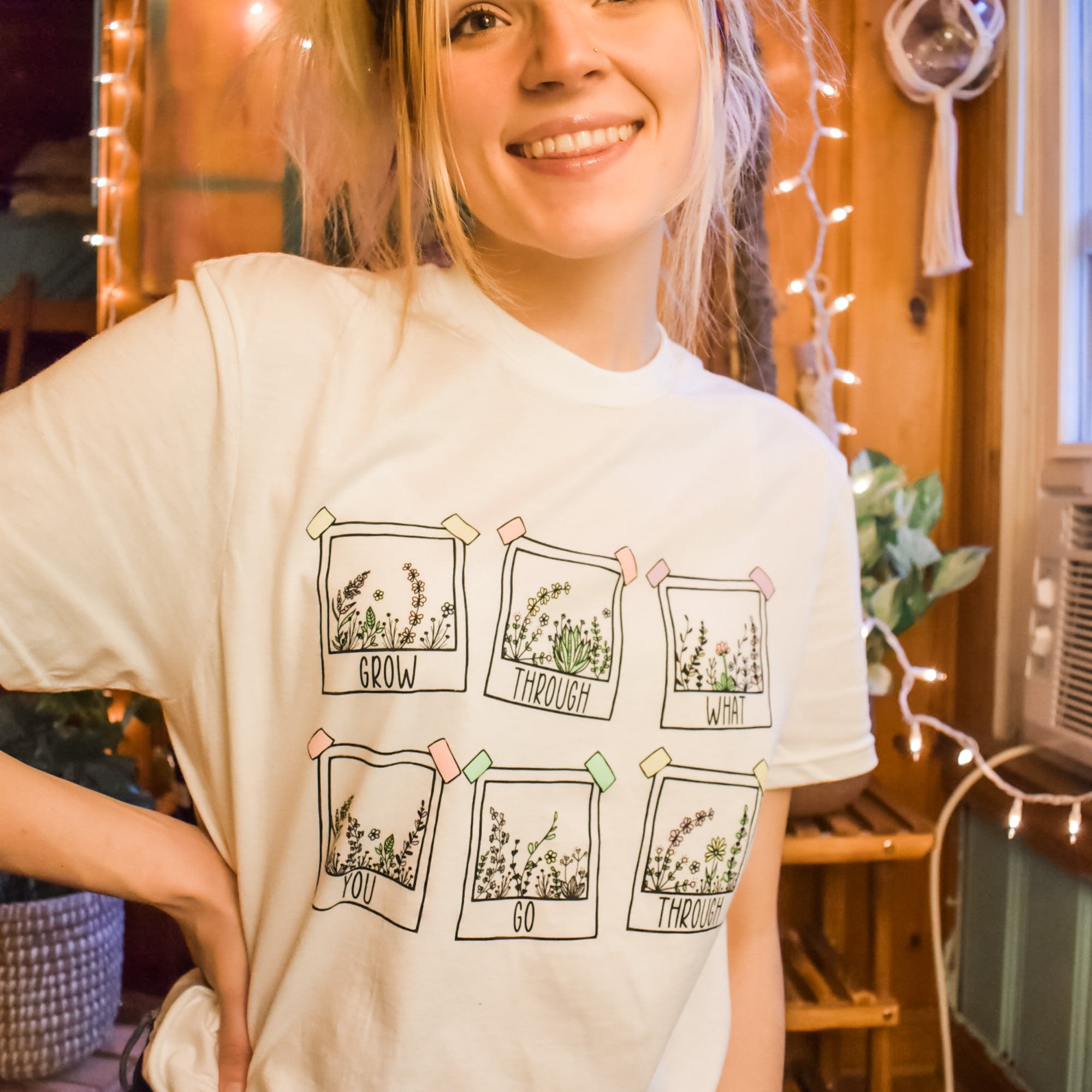 Grow Through What You Go Through positive message tee by Funnel Cake Tree cream unisex t-shirt featuring six floral polaroid-style frames and uplifting lettering, modeled indoors with string lights and a cozy vibe.