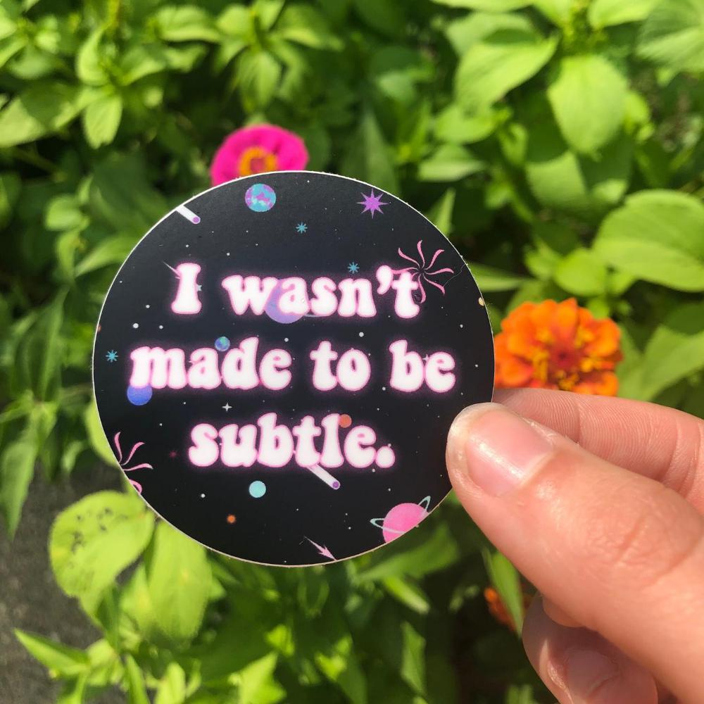 Round black sticker with space graphics held by a hand. Text reads: I wasn't made to be subtle.