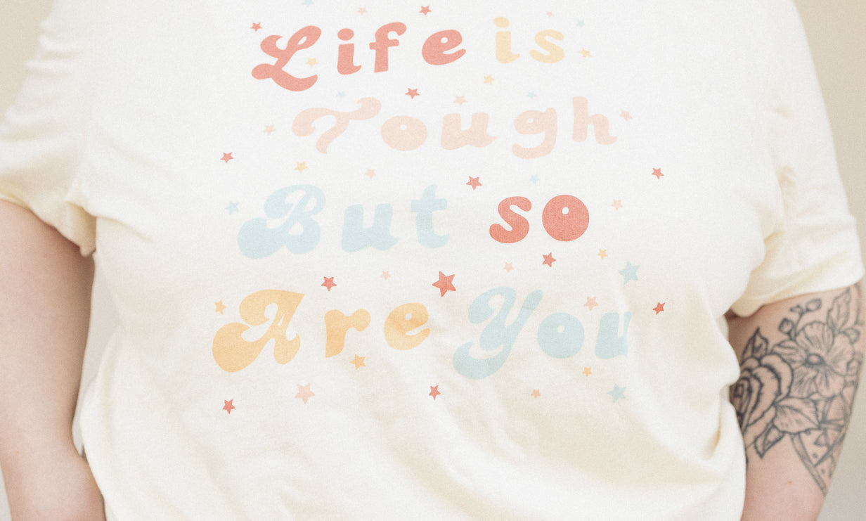 Close-up of cream ‘Life is Tough But So Are You’ graphic tee showing colorful retro typography and soft cotton texture.