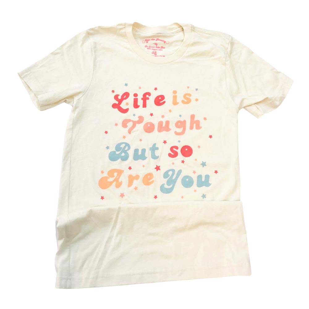 Flat lay of a cream ‘Life Is Tough But So Are You’ graphic tee with colorful retro text on a white background, neatly folded for product display.