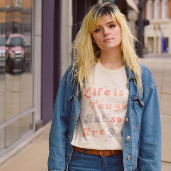 Woman wearing cream ‘Life is Tough But So Are You’ graphic tee styled with a denim jacket and jeans in an urban setting.