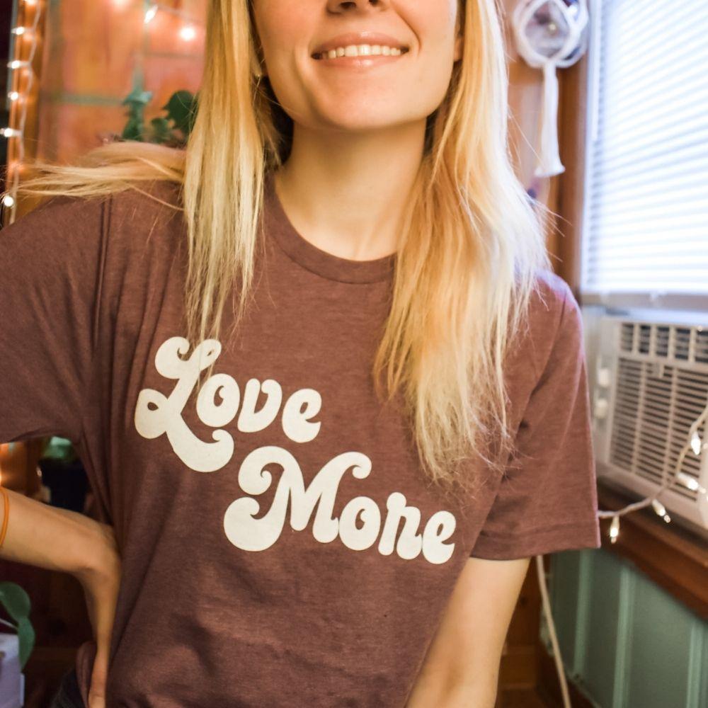 Woman wearing a burgundy ‘Love More’ graphic tee with bold white retro lettering, smiling indoors under warm cozy lighting, printed in the USA by Funnel Cake Tree.