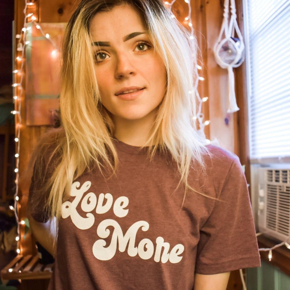 Woman wearing a burgundy ‘Love More’ graphic tee with bold white retro lettering, standing indoors surrounded by warm string lights, printed in the USA by Funnel Cake Tree.