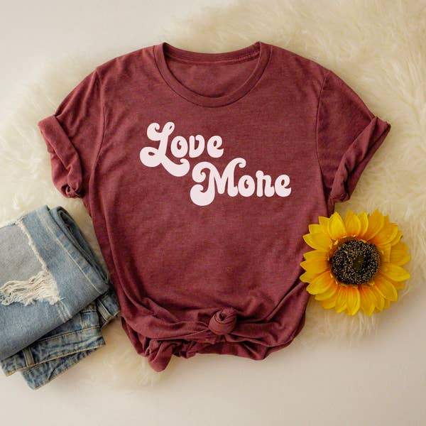 Flat lay of maroon ‘Love More’ graphic tee with bold white retro lettering, styled with denim jeans and a sunflower on a cozy cream background, printed in the USA by Funnel Cake Tree.