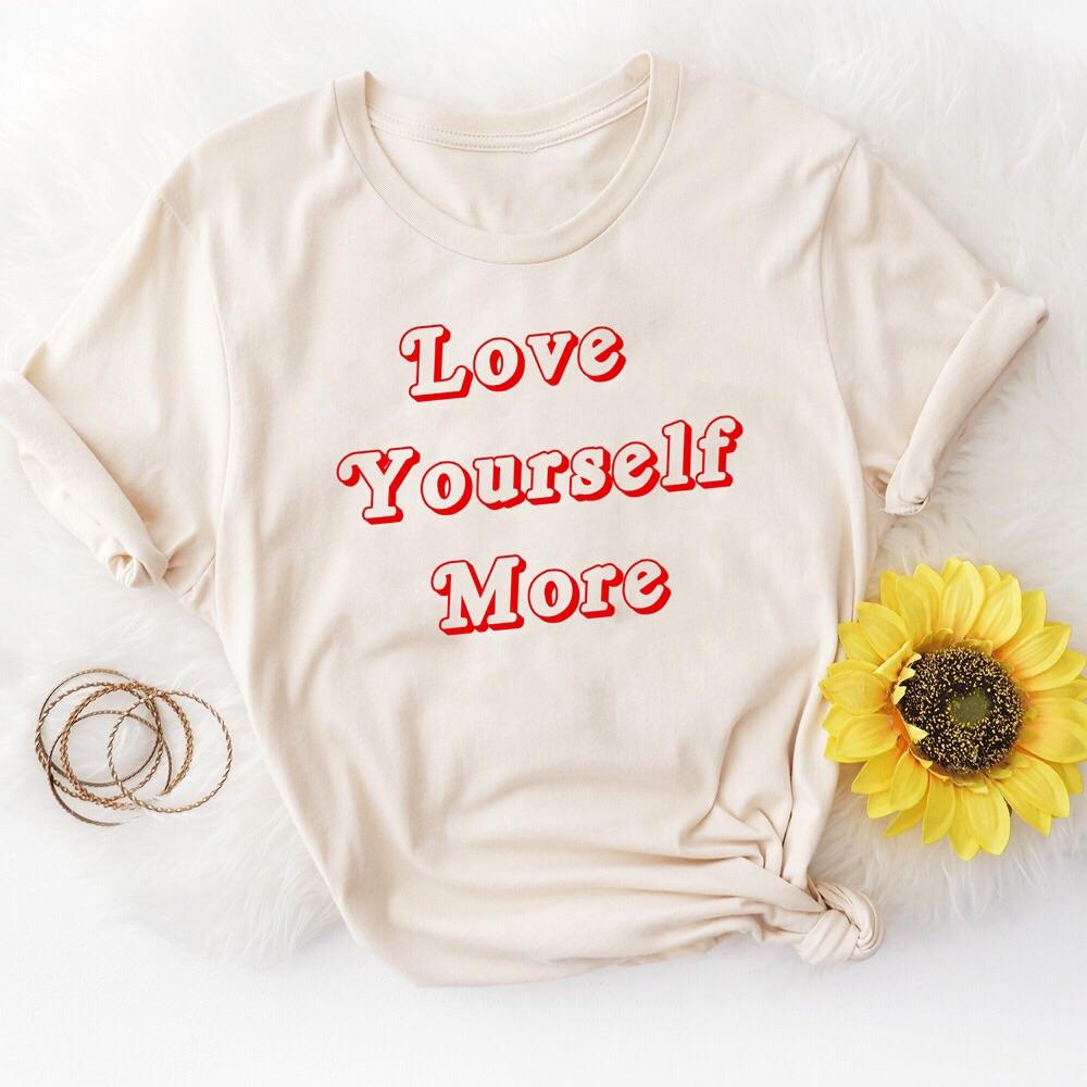 Beige t-shirt with red retro lettering that says ‘Love Yourself More’ styled on white fur background with a sunflower and bracelets.