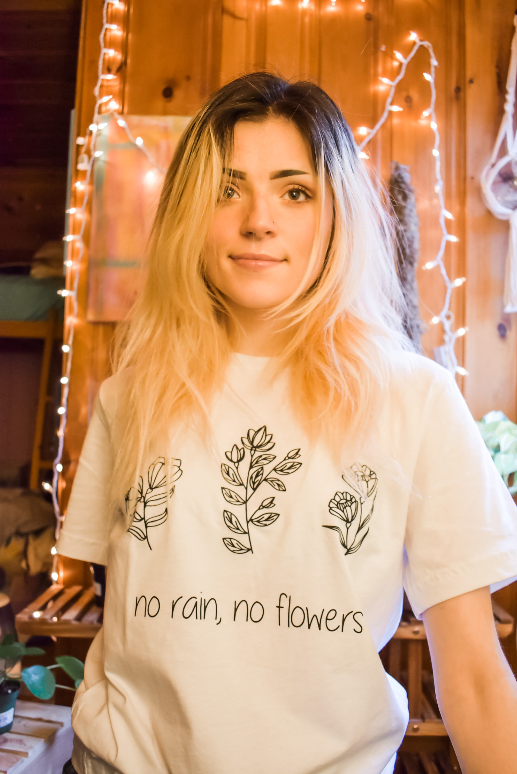 Woman wearing a white ‘No Rain, No Flowers’ t-shirt with minimalist black floral line art, standing in a cozy room with string lights.