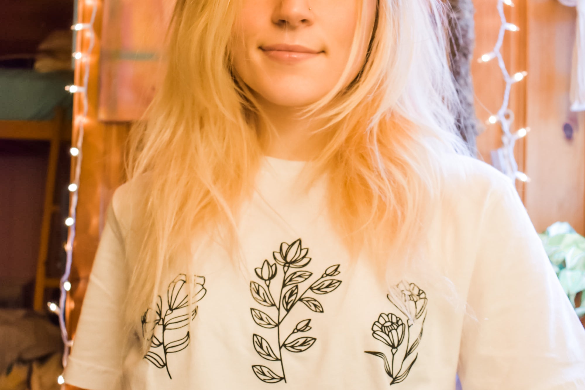 Woman wearing a white ‘No Rain, No Flowers’ t-shirt with minimalist black floral line art, standing in a cozy room with string lights.