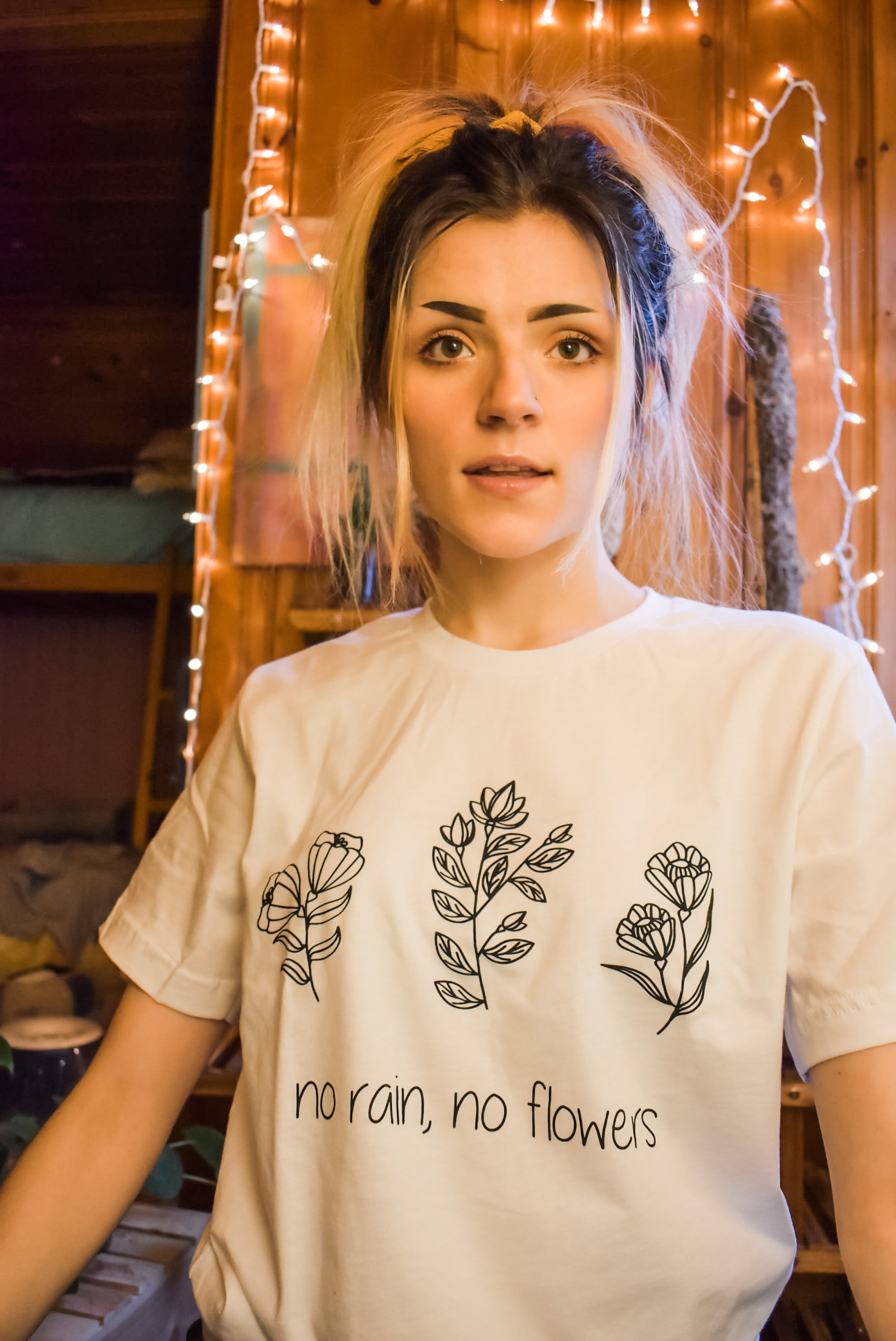 Woman wearing a white ‘No Rain, No Flowers’ t-shirt with minimalist black floral line art, standing in a cozy room with string lights.