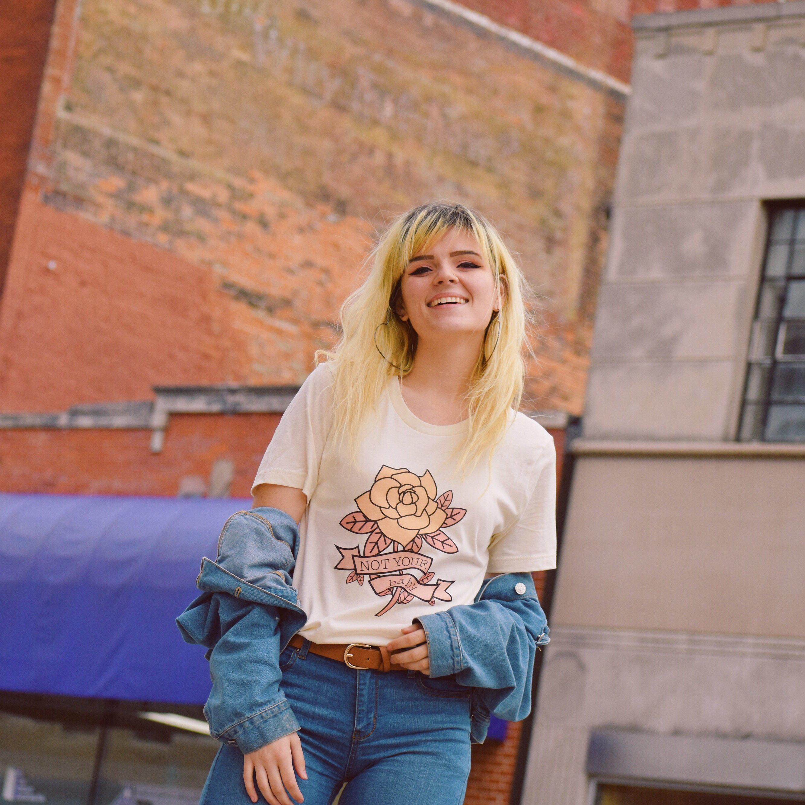 Woman wearing a cream ‘Not Your Babe’ graphic tee featuring a rose design and banner text, styled with a denim jacket and jeans against a city backdrop.
