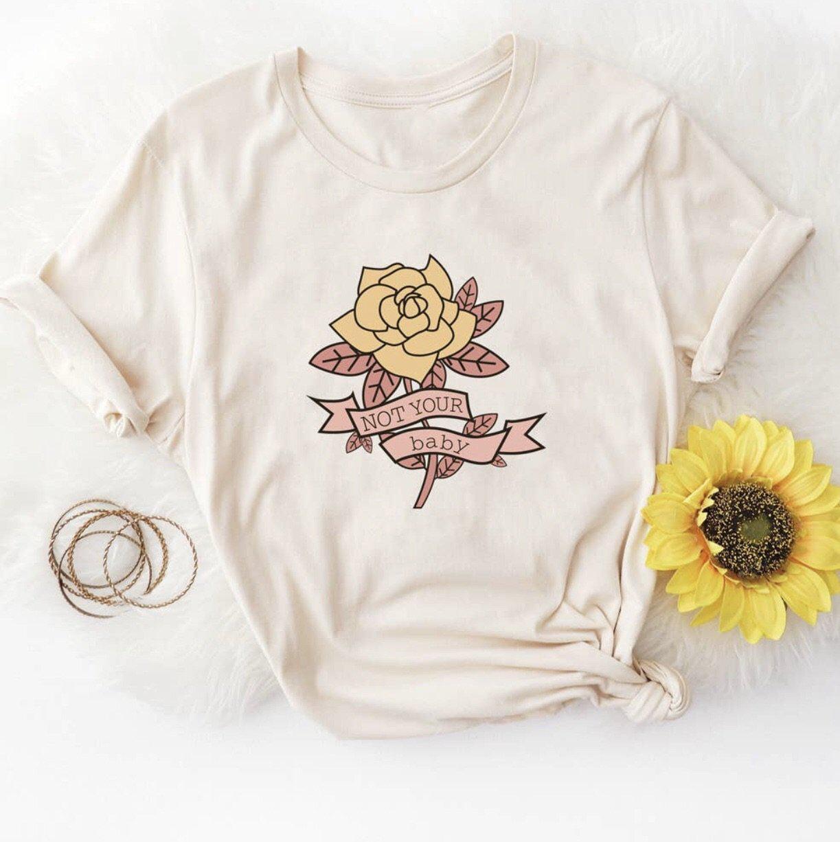 Flat lay of a beige ‘Not Your Baby’ graphic tee featuring a rose illustration and banner text, styled with a sunflower and gold bangles on a white background.