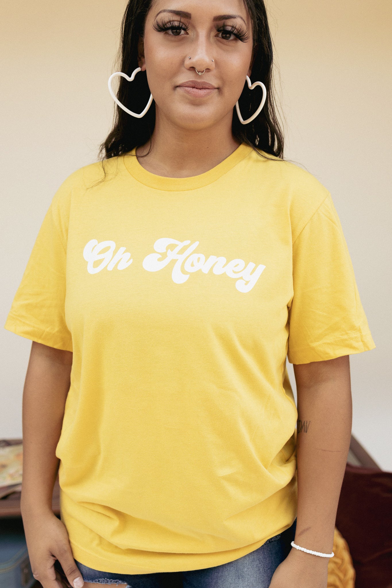 Woman wearing a yellow ‘Oh Honey’ graphic tee styled casually with denim jeans and heart-shaped earrings.