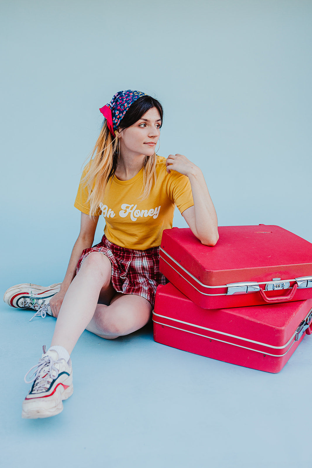 Woman wearing a yellow ‘Oh Honey’ graphic t-shirt styled with a red plaid skirt, vintage suitcases, and a floral headscarf against a light blue background.