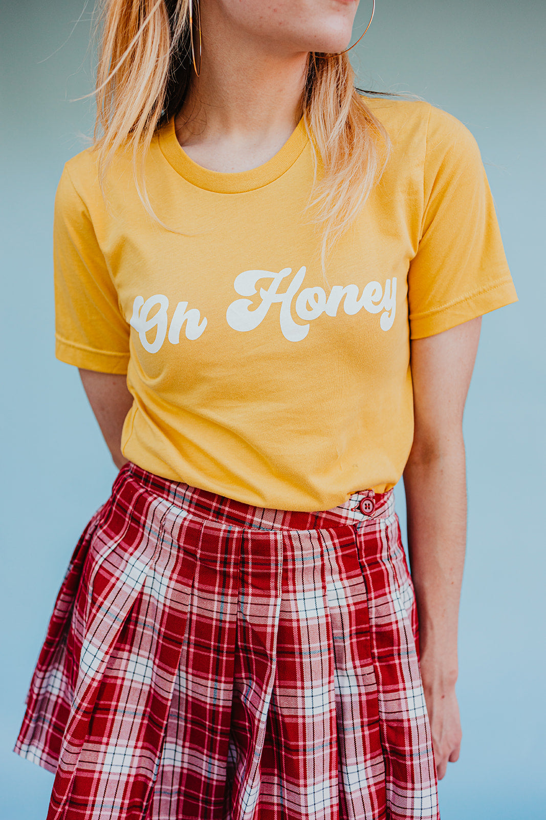 Close-up of a woman wearing a yellow ‘Oh Honey’ graphic tee paired with a red plaid skirt against a light blue background.