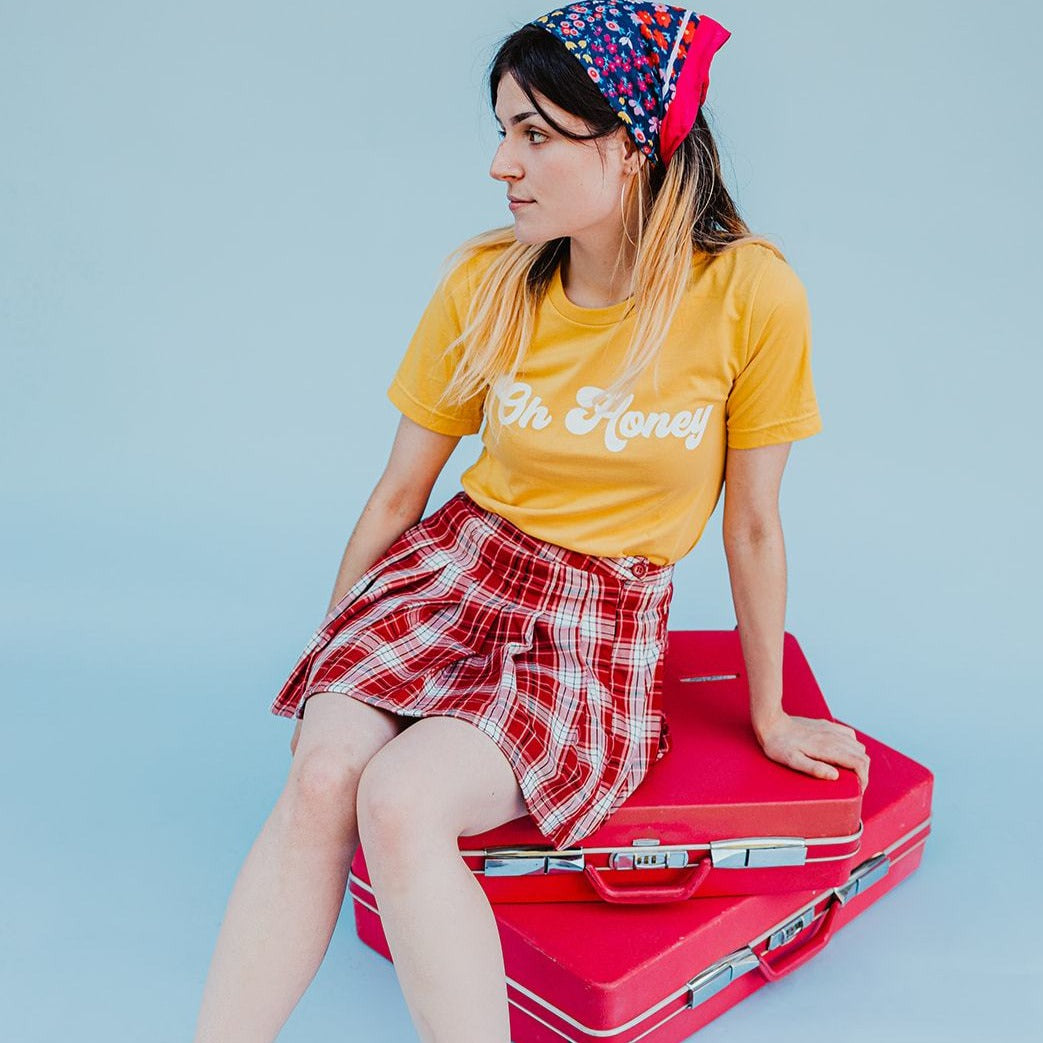 Woman wearing a yellow ‘Oh Honey’ graphic tee with a red plaid skirt, sitting on vintage red suitcases against a blue background.
