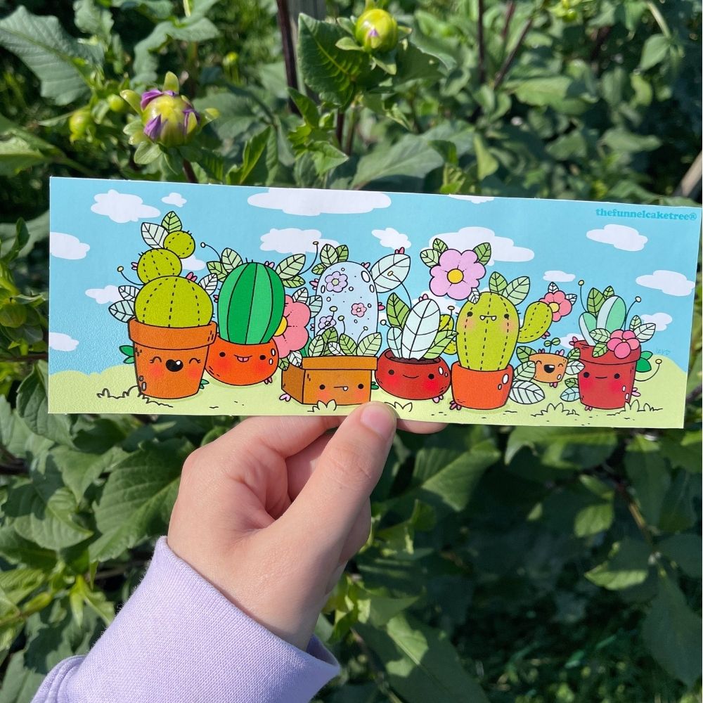 Rectangular sticker with cartoon cacti in smiling pots against a blue sky, held in front of green leaves.