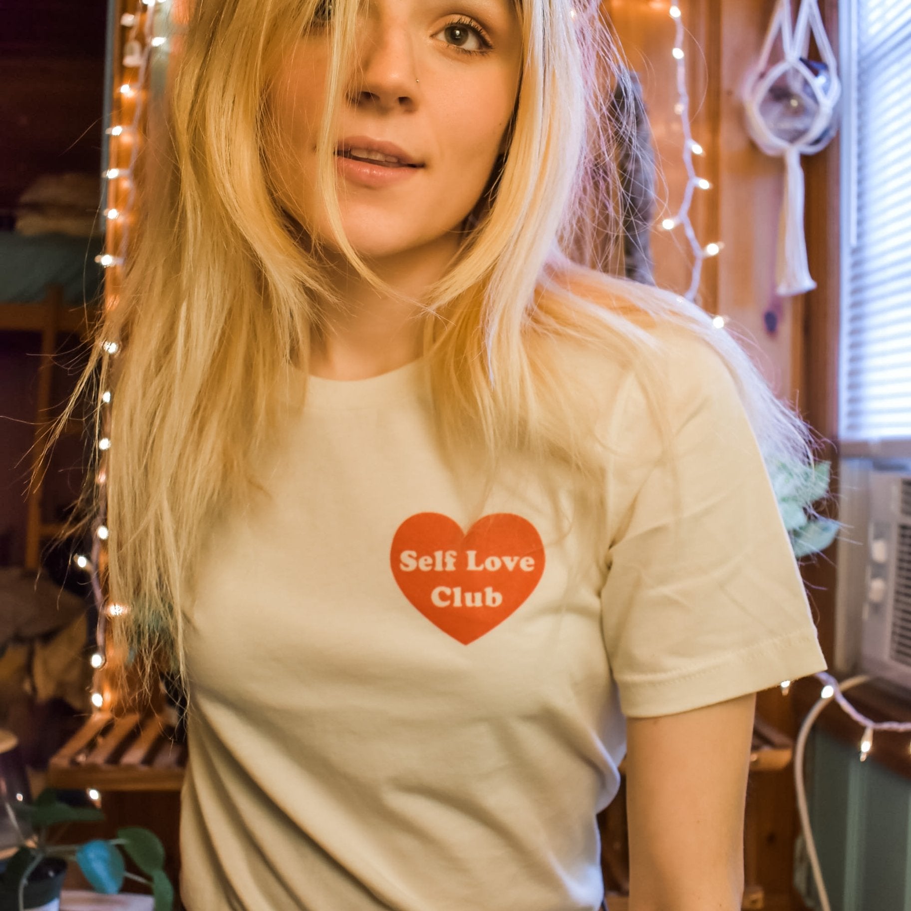 Woman wearing a cream Self Love Club graphic tee with a red heart print, smiling indoors with cozy string lights in the background.