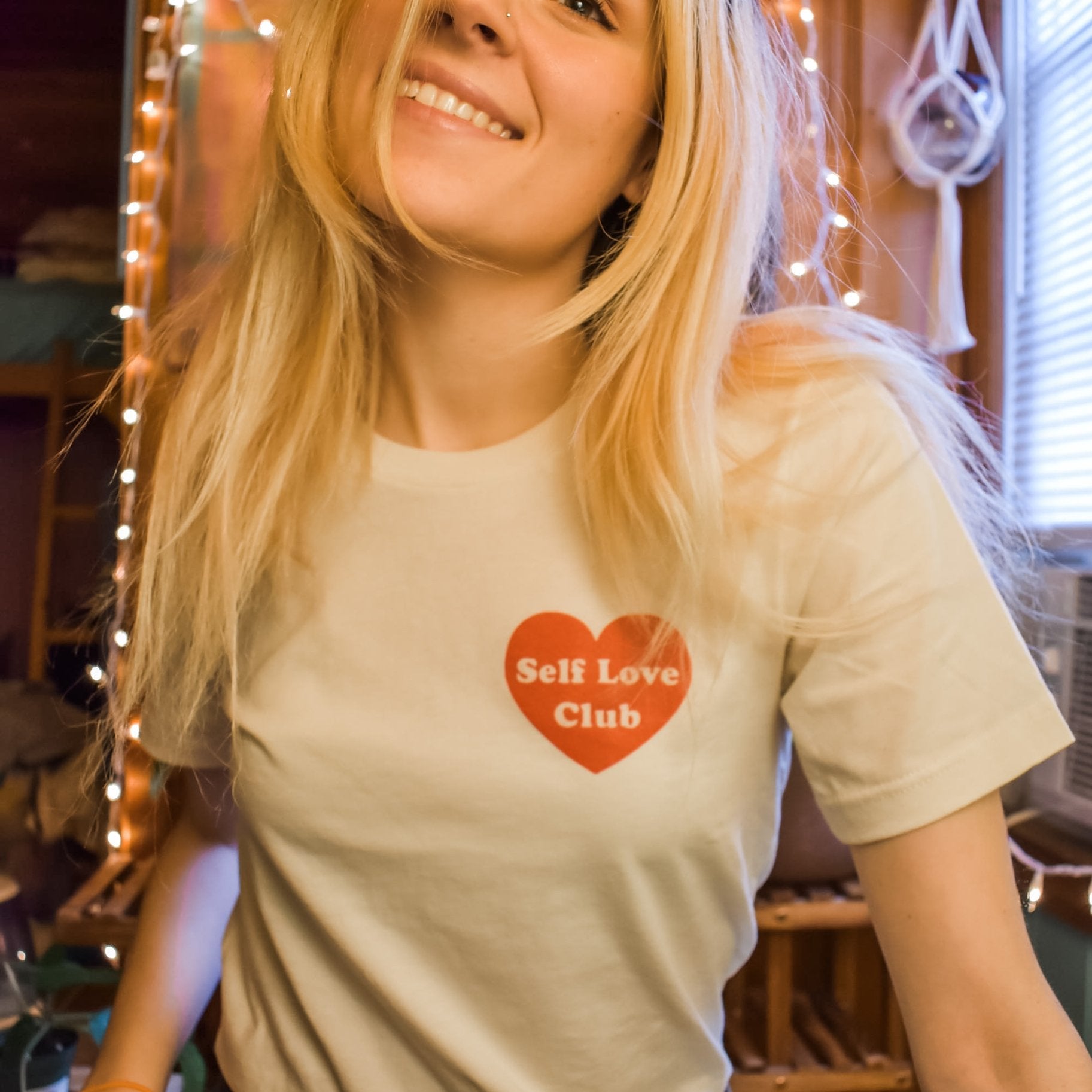 Woman smiling while wearing a cream Self Love Club graphic tee with a red heart design, standing in a cozy room with string lights.