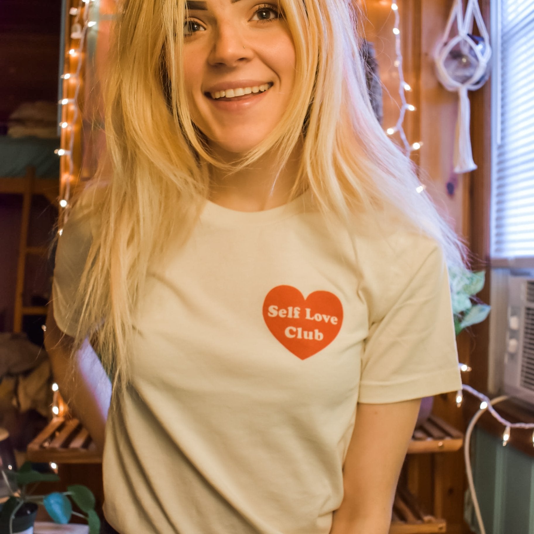 Woman wearing a cream Self Love Club graphic tee with a red heart design, standing indoors with warm string lights in the background.