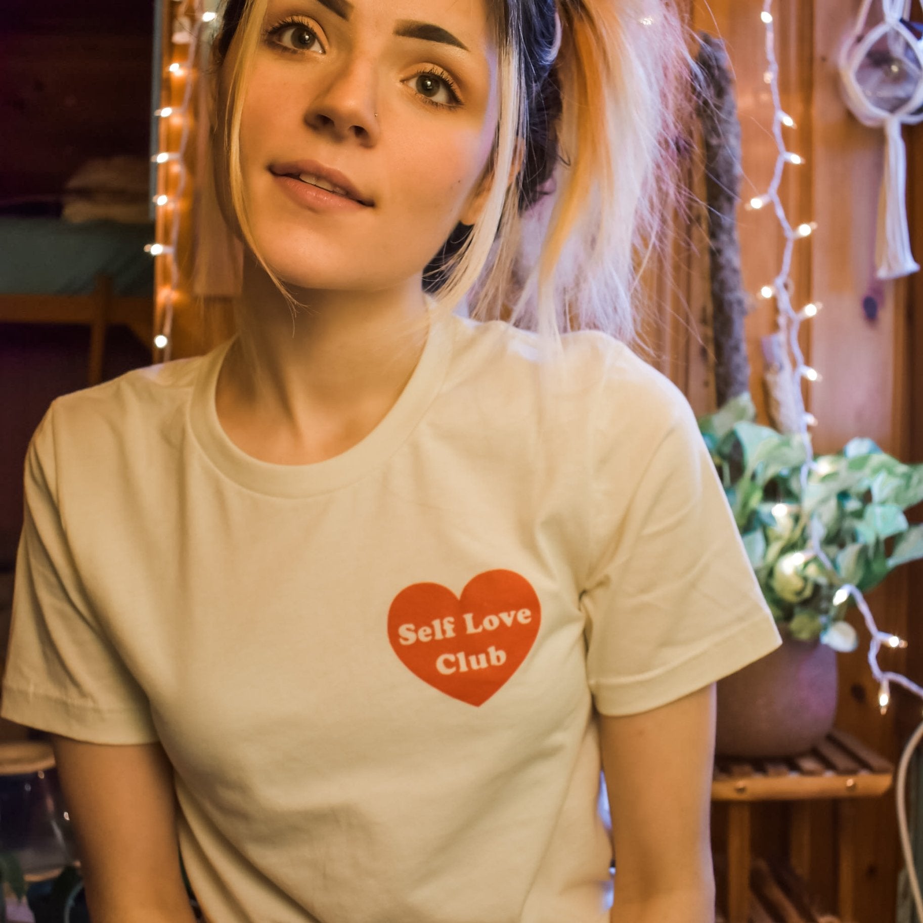 Woman wearing a cream Self Love Club graphic tee with a red heart design, sitting in a cozy wooden room with soft lighting and string lights.