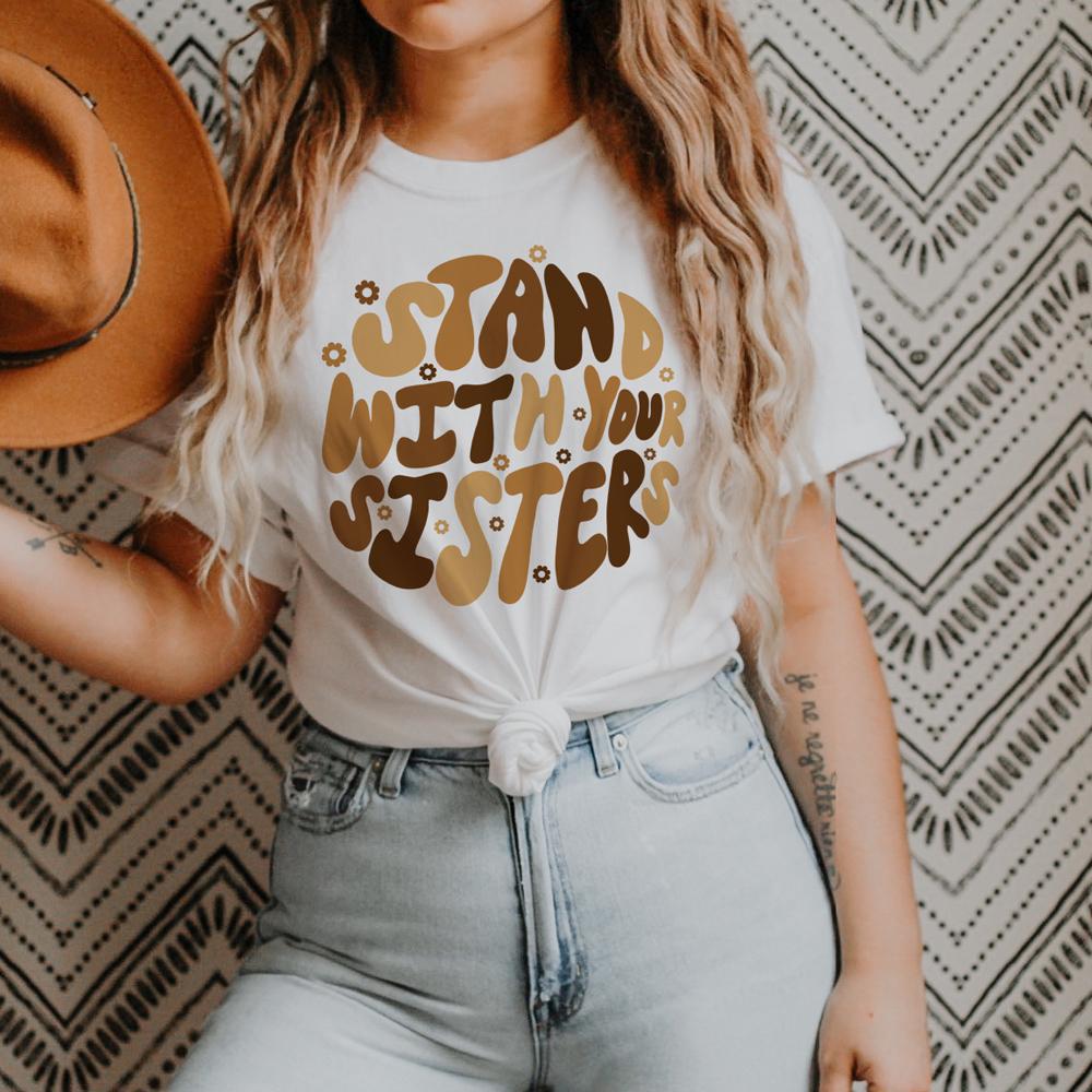 Stand With Your Sisters lifestyle graphic tee by Funnel Cake Tree , white unisex t-shirt with retro brown lettering and daisy accents, styled with light-wash jeans and a hat against a boho patterned background.